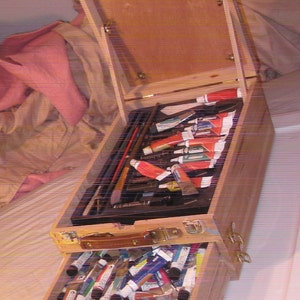 May include: Wooden artist's paint box with a hinged lid and two pull-out drawers. The top drawer contains paint tubes and brushes. The bottom drawer is filled with paint tubes. The box is made of light-colored wood.