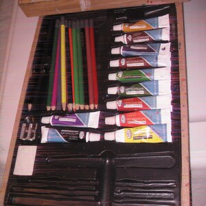 May include: A wooden artist's box with an open drawer revealing art supplies. The box contains watercolor tubes, colored pencils, a pencil sharpener, and other painting tools. The watercolor tubes are in various colors, including blue, red, and yellow.