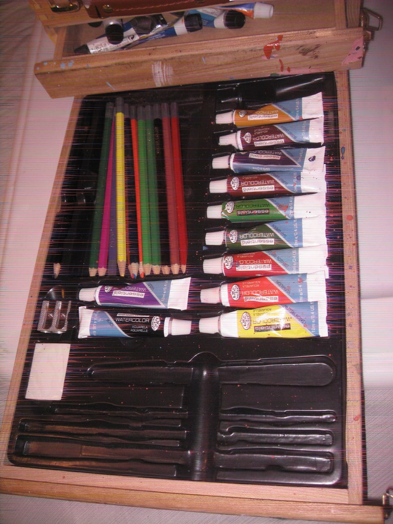 May include: A wooden artist's box with an open drawer revealing art supplies. The box contains watercolor tubes, colored pencils, a pencil sharpener, and other painting tools. The watercolor tubes are in various colors, including blue, red, and yellow.