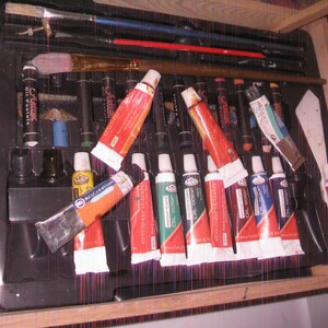 May include: A black art supply case filled with oil paints, brushes, and other painting tools. The case contains tubes of oil paint in various colors, including red, green, and blue. Paintbrushes of different sizes and a palette knife are also visible.