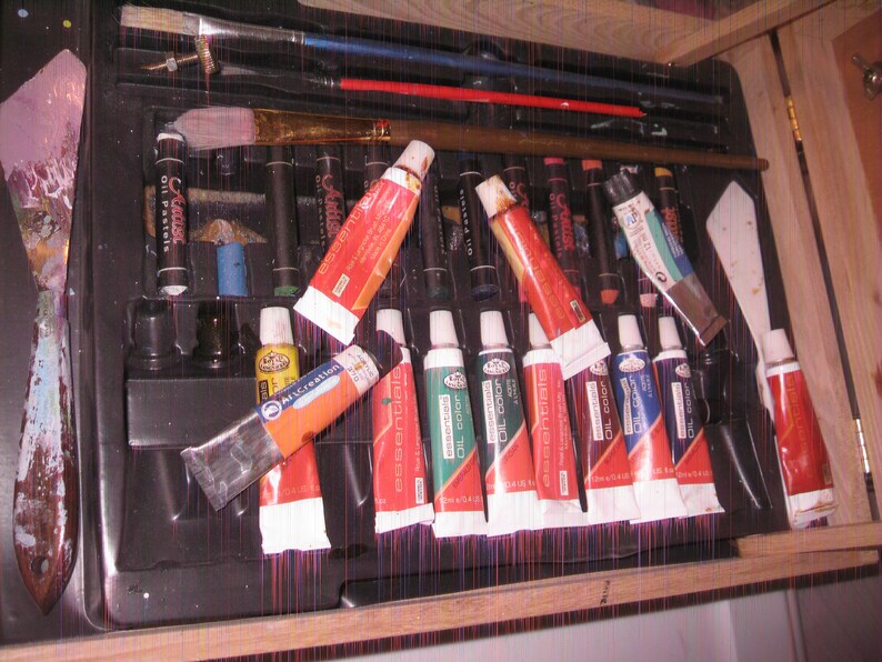 May include: A black art supply case filled with oil paints, brushes, and other painting tools. The case contains tubes of oil paint in various colors, including red, green, and blue. Paintbrushes of different sizes and a palette knife are also visible.