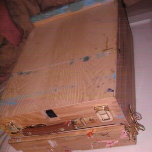 May include: A wooden artist's easel box, stained with paint splatters in various colors. The box has a brown leather handle, metal clasps, and a blue-painted edge. The box is designed to hold art supplies.
