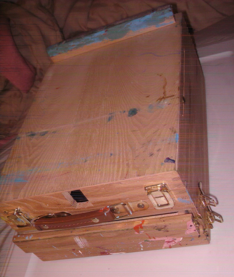 May include: A wooden artist's easel box, stained with paint splatters in various colors. The box has a brown leather handle, metal clasps, and a blue-painted edge. The box is designed to hold art supplies.