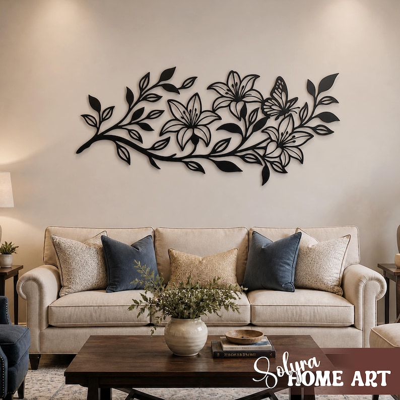 Puede incluir: Arte mural floral de metal negro con lirios y una mariposa. La pieza decorativa est&aacute; montada en una pared de color neutro sobre un sof&aacute; beige con cojines azules y beige. El arte a&ntilde;ade un toque de elegancia inspirada en la naturaleza a la sala de estar.
