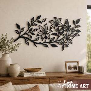 Puede incluir: Arte mural floral de metal negro con lirios y una mariposa. La pieza decorativa est&aacute; montada en una pared de color neutro sobre un mueble de madera. El mueble contiene libros, jarrones y una foto enmarcada.