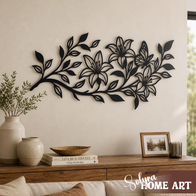 Puede incluir: Arte mural floral de metal negro con lirios y una mariposa. La pieza decorativa est&aacute; montada en una pared de color neutro sobre un mueble de madera. El mueble contiene libros, jarrones y una foto enmarcada.