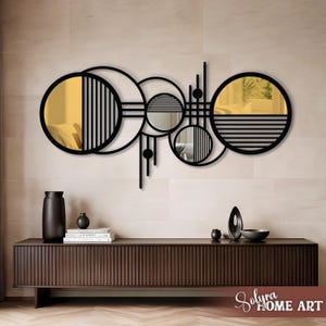 May include: A modern wall mirror with a geometric design. The mirror features black metal circles, lines, and semi-circles, with gold and reflective mirror surfaces. The design includes vertical and horizontal lines, and small black spheres. The mirror is mounted on a light-colored wall.