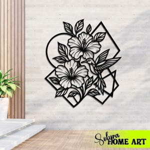 May include: Black metal wall art featuring hibiscus flowers, leaves, a hummingbird, and geometric shapes. The artwork is mounted on a light gray textured wall. The bottom right corner has the text "Solyra HOME ART".