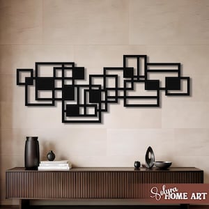 May include: Black metal wall art with an abstract geometric design of interconnected squares and rectangles. The artwork is mounted on a neutral-toned wall above a dark wood cabinet with decorative items. The Solyra Home Art logo is visible.