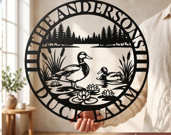 May include: A circular black metal wall sign featuring a laser-cut design of two ducks in a pond with water lilies, cattails, and a forest background. The text THE ANDERSONS and DUCK FARM is cut into the circular border of the metal sign.