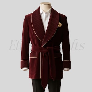 May include: A burgundy velvet smoking jacket with a quilted shawl collar and gold trim. The jacket features a matching belt, pocket details, and cuffs. A white dress shirt and a gold pocket square are visible.
