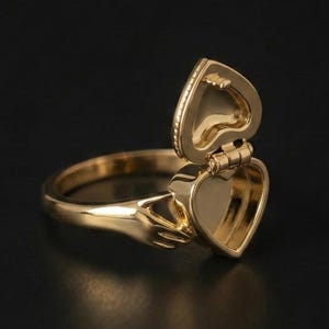 Könnte beinhalten: Ein goldfarbener Metallring mit einem aufklappbaren, herzförmigen Medaillon. Die Ringschiene ist so gestaltet, dass zwei Hände die Basis des herzförmigen Medaillons halten, welches in geöffneter Position einen hohlen Innenraum zeigt.