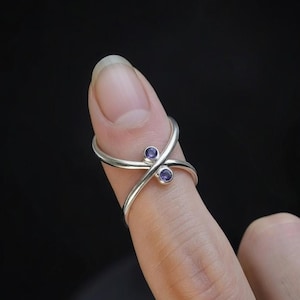 May include: A silver ring with a unique crisscross design, featuring two small, round, purple gemstones. The ring is worn on a finger, showcasing its elegant and minimalist style. The gemstones are set in the center of the ring's design.