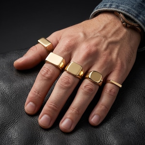 Könnte beinhalten: Ein Set aus fünf goldfarbenen Ringen an den Fingern. Die Ringe haben verschiedene Formen, darunter rechteckige, quadratische und abgerundete Siegelring-Stile sowie ein dünnes, flaches Band. Jeder Ring hat eine glatte, polierte Metalloberfläche.