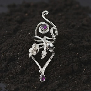 May include: A silver-toned ring with an intricate floral design. The ring features a flower, leaves, and a swirling pattern, with a small, round, purple gemstone at the top and bottom. The ring is set against a dark background.