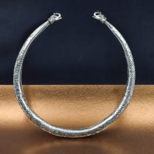 May include: A silver-toned necklace with intricate floral and scrollwork patterns. The ends of the necklace are sculpted into stylized animal heads. The necklace is displayed against a two-tone background of navy blue and gold.
