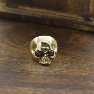May include: A gold-coloured skull ring featuring two red gemstones as eyes. The ring is made of a shiny metal and is set against a wooden background. The skull design is detailed and the gemstones add a pop of colour.