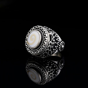 Op de afbeelding: Een zilveren ring met een ovale, witte steen met een gouden spiraalontwerp. De ring heeft een sierlijk, gedetailleerd patroon rond de steen en op de band. De ring staat tegen een zwarte achtergrond, met zijn weerspiegeling zichtbaar.