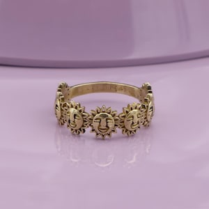 May include: A gold-colored ring featuring a repeating sun face design. The ring has a band with a series of sun faces around the circumference. The sun faces have smiling expressions and are set against a light purple background.