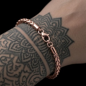 May include: A rose gold wheat chain bracelet featuring a polished finish and a secure lobster claw clasp. The bracelet is constructed with interlocking woven links that create a rounded, textured appearance, providing a consistent metallic sheen.