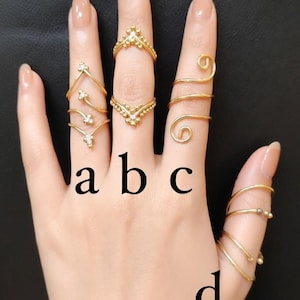Può includere: Una mano mostra vari anelli color oro. Gli stili includono fasce delicate, motivi floreali e avvolgimenti a spirale. Gli anelli sono etichettati a, b, c e d. La mano ha unghie smaltate.
