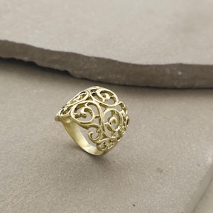 May include: A gold-coloured ring with an openwork design featuring swirling, ornate patterns. The ring has a rounded shape and a band that tapers towards the back. The intricate details create a delicate and elegant aesthetic, suitable for jewellery.