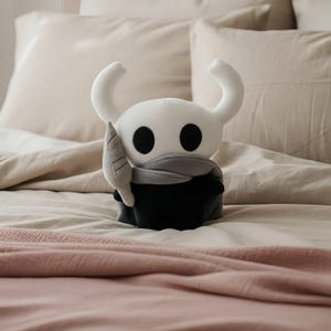 May include: A plush toy of a character with a white head, large black eyes, and curved horns. The character wears a gray scarf and a black cloak, holding a gray leaf-shaped object. The toy is on a bed with beige and pink bedding.