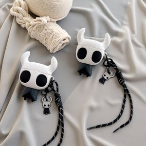 Puede incluir: Dos fundas para AirPod blancas y negras con temática de Hollow Knight con cordones negros y blancos. Las fundas presentan un diseño de personaje estilizado con grandes ojos negros y una pequeña capa. Un gorro de punto está en el fondo.