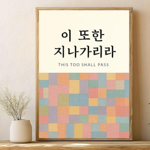 Puede incluir: Arte mural enmarcado con texto coreano y la traducción al inglés "This Too Shall Pass". La obra de arte presenta una sección superior color crema con caracteres coreanos negros y un patrón geométrico colorido debajo.