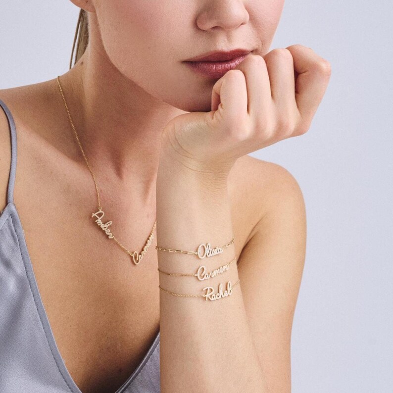 Puede incluir: Un collar y pulseras doradas con nombres personalizados. El collar dice "Amber" y "Caroline". Las pulseras dicen "Olivia", "Carmen" y "Rachel". Las joyas se muestran en el cuello y la mu&ntilde;eca de una persona.