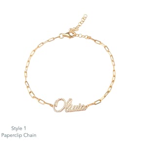 Puede incluir: Pulsera de cadena de clip de papel dorada con el nombre "Olivia" escrito con peque&ntilde;os cristales transparentes. La pulsera tiene un cierre de mosquet&oacute;n. El texto "Style 1 Paperclip Chain" est&aacute; en la parte inferior.