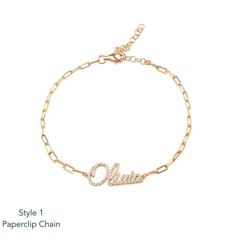 Puede incluir: Pulsera de cadena de clip de papel dorada con el nombre "Olivia" escrito con peque&ntilde;os cristales transparentes. La pulsera tiene un cierre de mosquet&oacute;n. El texto "Style 1 Paperclip Chain" est&aacute; en la parte inferior.