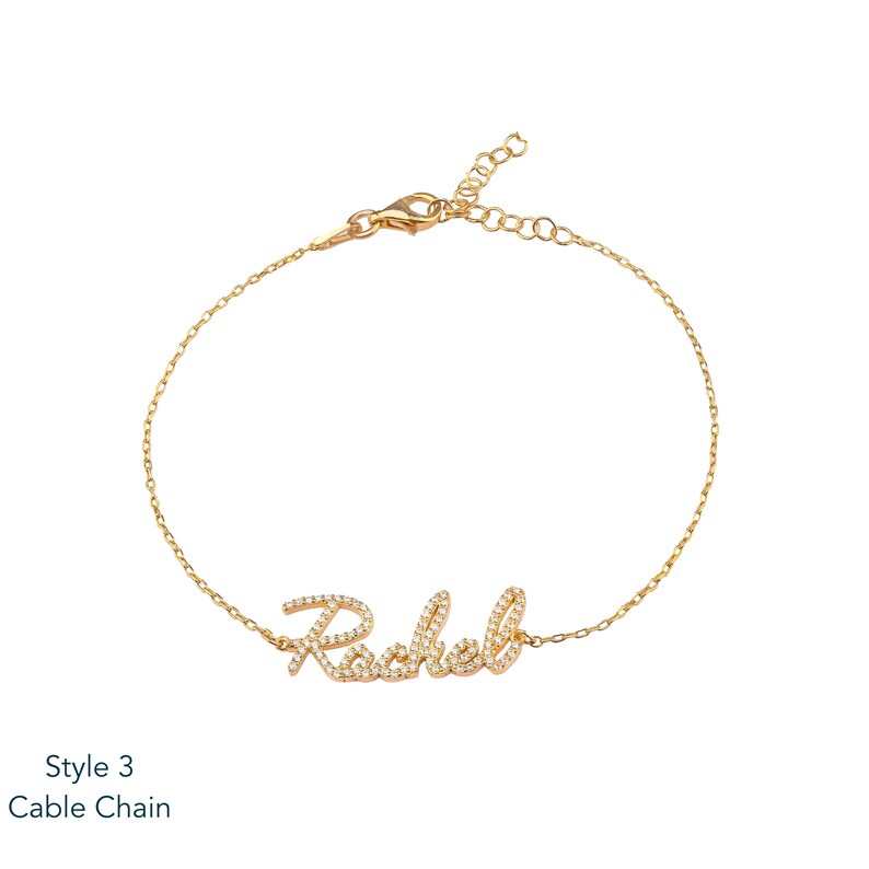 Puede incluir: Una pulsera de cadena dorada con el nombre "Rachel" escrito en cursiva, incrustado con peque&ntilde;as piedras preciosas transparentes. La pulsera tiene un cierre de mosquet&oacute;n y una cadena ajustable. El texto "Style 3 Cable Chain" est&aacute; en la esquina inferior izquierda.