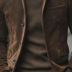 May include: A brown suede blazer with three buttons and a chest pocket. The blazer is worn over a brown knit sweater. The overall look is a classic, casual style.
