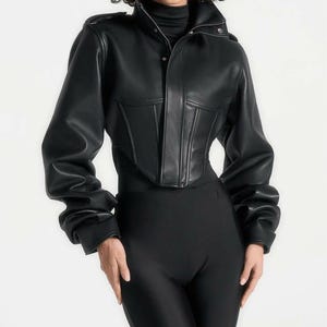 May include: Black cropped leather jacket with a high collar and button closure. The jacket has a corset-style bodice and long sleeves. The model is wearing black leggings and a black turtleneck.