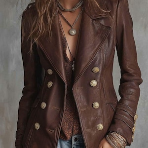 Peut inclure: Veste en cuir marron avec des boutons dorés. La veste a une conception croisée et un col cranté. Les manches ont des détails de boutons dorés décoratifs. Le modèle porte des colliers superposés et des bracelets.