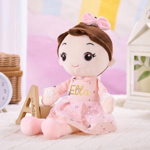 May include: A soft doll with brown hair and a pink bow. The doll is dressed in a pink dress with the name "Ella" in gold lettering. The doll is sitting on a white surface.