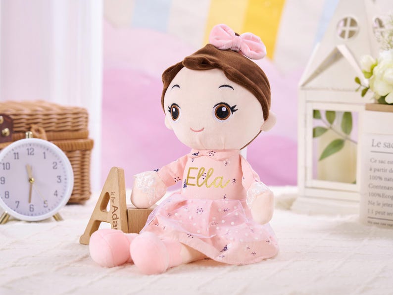May include: A soft doll with brown hair and a pink bow. The doll is dressed in a pink dress with the name "Ella" in gold lettering. The doll is sitting on a white surface.