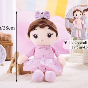 May include: A soft doll with brown hair and a lavender bow, dressed in a pink dress with a dotted skirt. The doll is 28 cm tall and 45 cm overall. A smaller image shows two similar dolls.