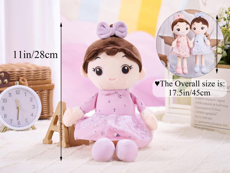 May include: A soft doll with brown hair and a lavender bow, dressed in a pink dress with a dotted skirt. The doll is 28 cm tall and 45 cm overall. A smaller image shows two similar dolls.