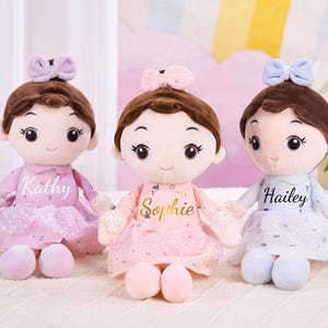 May include: Three soft dolls with brown hair, bows, and dresses in pink and blue. Each doll has a name embroidered on the front: Kathy, Sophie, and Hailey. The dolls are designed to look like little girls.