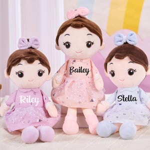 May include: Three plush dolls with brown hair and bows, each wearing a dress with a name printed on it: Riley, Bailey, and Stella. The dolls have large eyes and are sitting on a white surface.