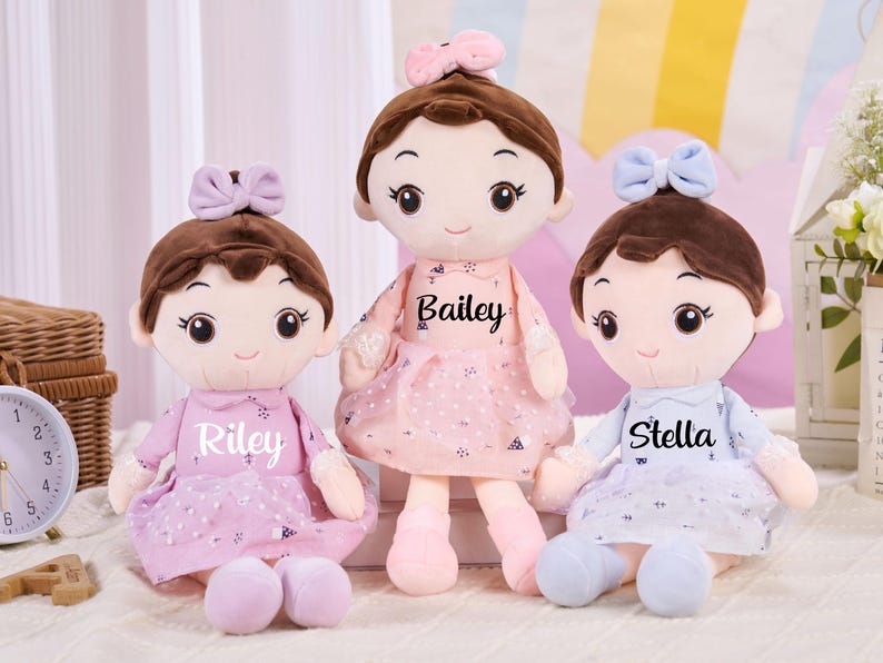 May include: Three plush dolls with brown hair and bows, each wearing a dress with a name printed on it: Riley, Bailey, and Stella. The dolls have large eyes and are sitting on a white surface.