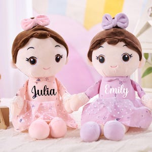 May include: Two plush dolls with brown hair and bows, wearing pink dresses. One doll has "Julia" printed on the dress, the other has "Emily". Each doll holds a pair of matching coloured plush balls. The dolls are on a white surface.