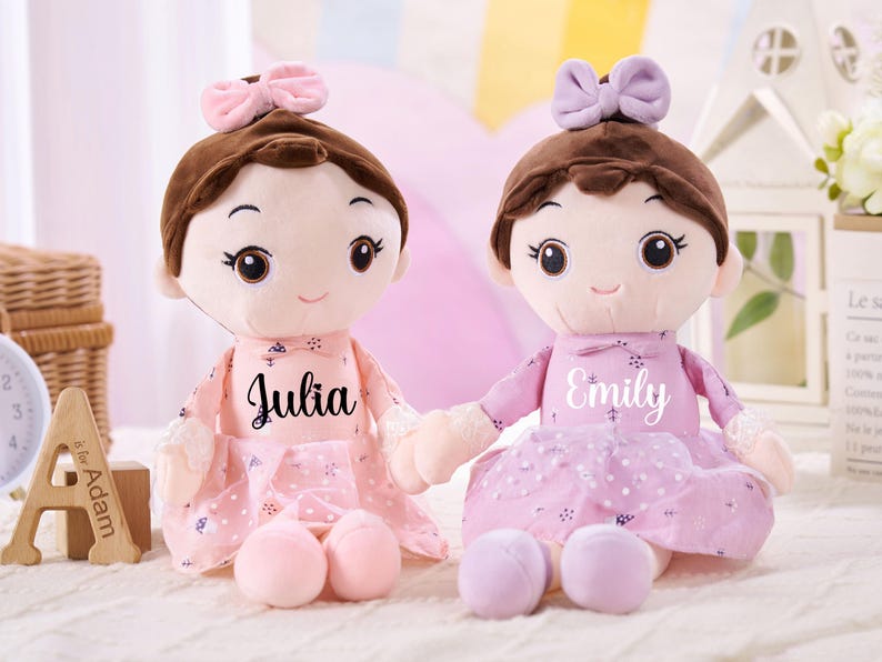 May include: Two plush dolls with brown hair and bows, wearing pink dresses. One doll has "Julia" printed on the dress, the other has "Emily". Each doll holds a pair of matching coloured plush balls. The dolls are on a white surface.