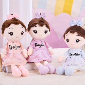 May include: Three soft dolls with brown hair and bows, each wearing a dress and personalised with a name: Evelyn, Kayla, and Sophia. The dolls have large eyes and are in shades of pink and blue.