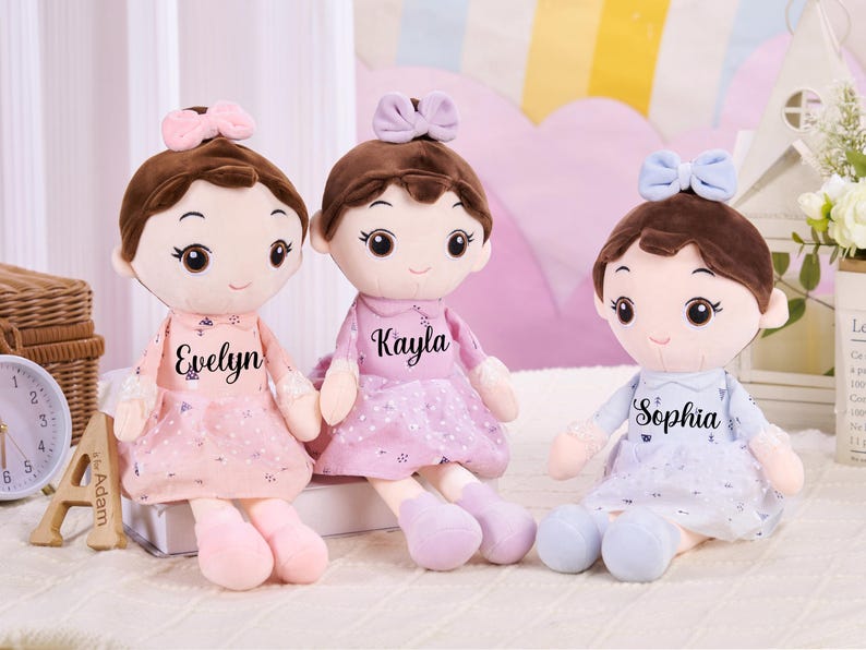 May include: Three soft dolls with brown hair and bows, each wearing a dress and personalised with a name: Evelyn, Kayla, and Sophia. The dolls have large eyes and are in shades of pink and blue.