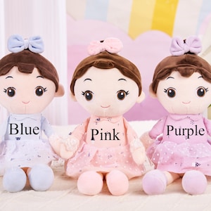 May include: Three plush dolls in blue, pink, and purple dresses, each with a matching bow and the colour name printed on the dress. The dolls have brown hair and are sitting on a white surface.