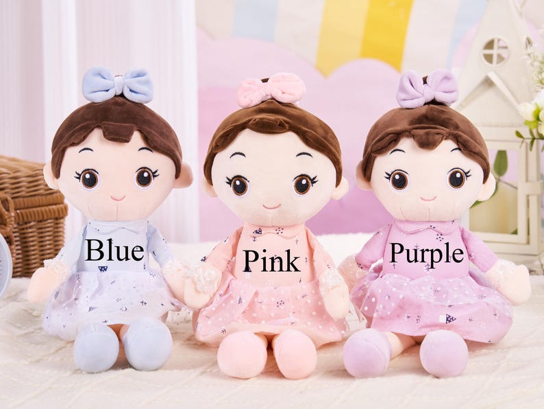 May include: Three plush dolls in blue, pink, and purple dresses, each with a matching bow and the colour name printed on the dress. The dolls have brown hair and are sitting on a white surface.