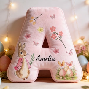 Peut inclure: Un coussin en forme de lettre "A" rose, orné d'un lapin, de papillons et de motifs floraux. Le nom "Amelia" est imprimé en noir. Décorations de Pâques avec des œufs et une figurine de lapin.